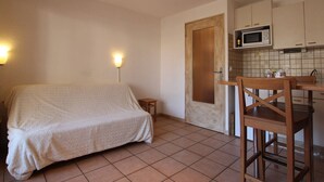 Una habitación pequeña y funcional con cama, silla, mesa y kitchenette.