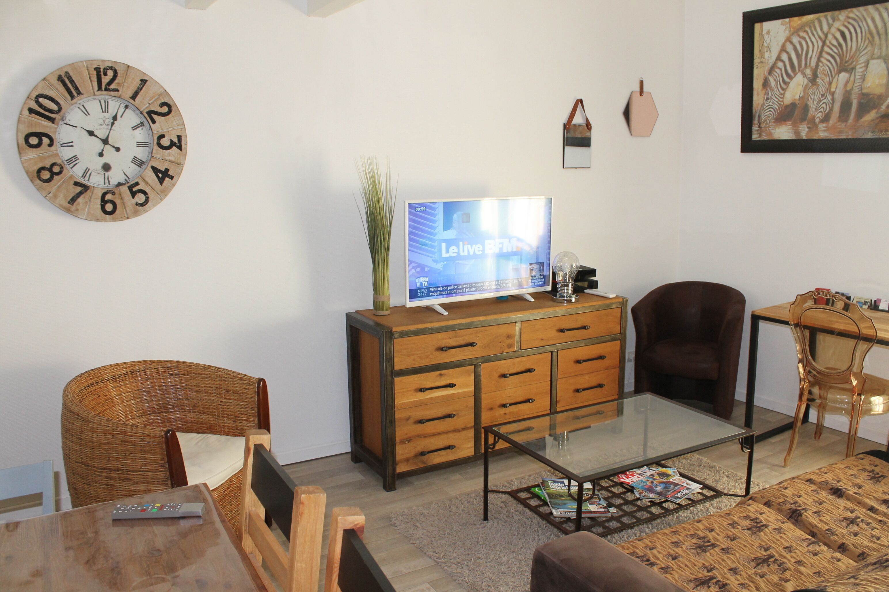 Een woonkamer met een houten kast, een televisie, een rieten fauteuil, een glazen salontafel en een ingelijst kunstwerk aan de muur.