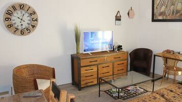 Een woonkamer met een houten kast, een televisie, een rieten fauteuil, een glazen salontafel en een ingelijst kunstwerk aan de muur.