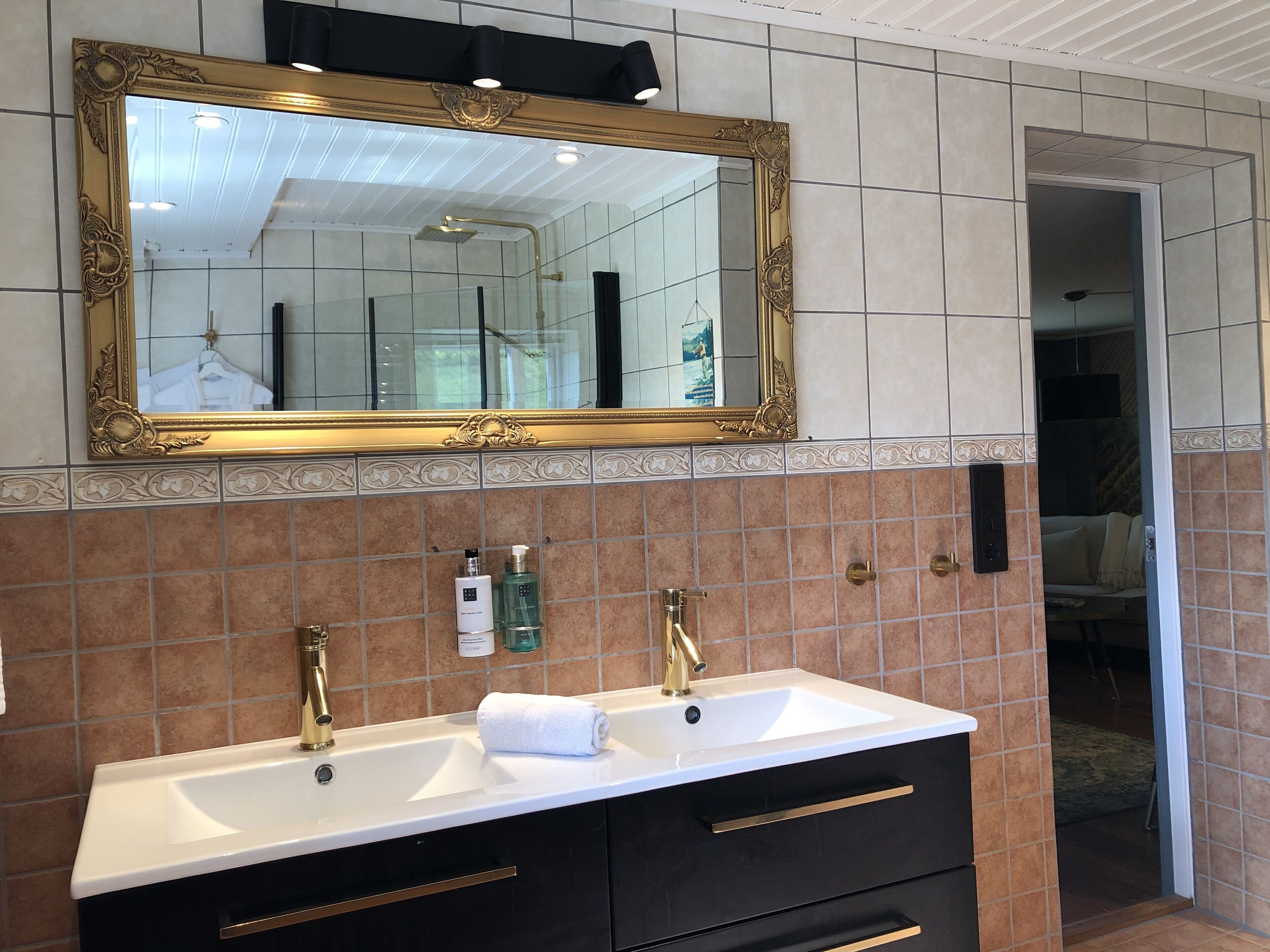 Un baño con un espejo grande y ornamentado, lavamanos dobles con grifos dorados y una toalla sobre la encimera.