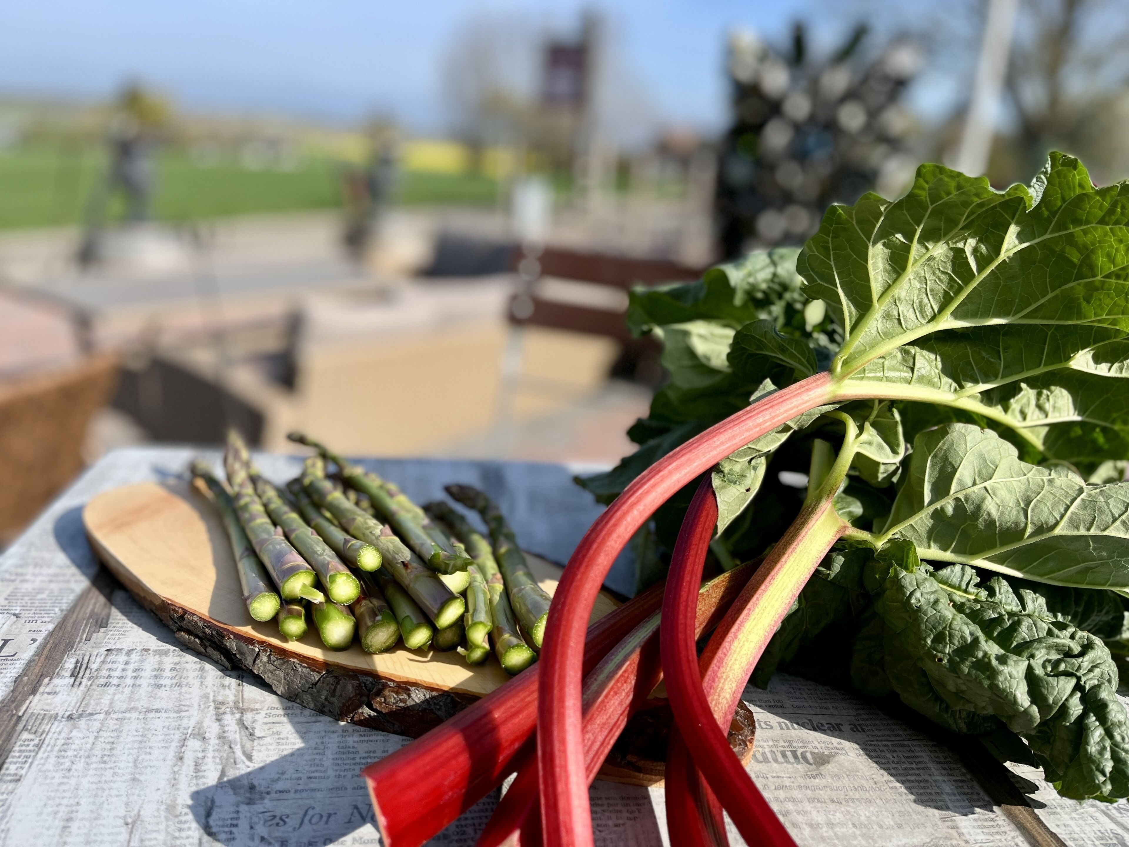 A wooden table with vegetables, including asparagus and leafy greens, on a newspaper.