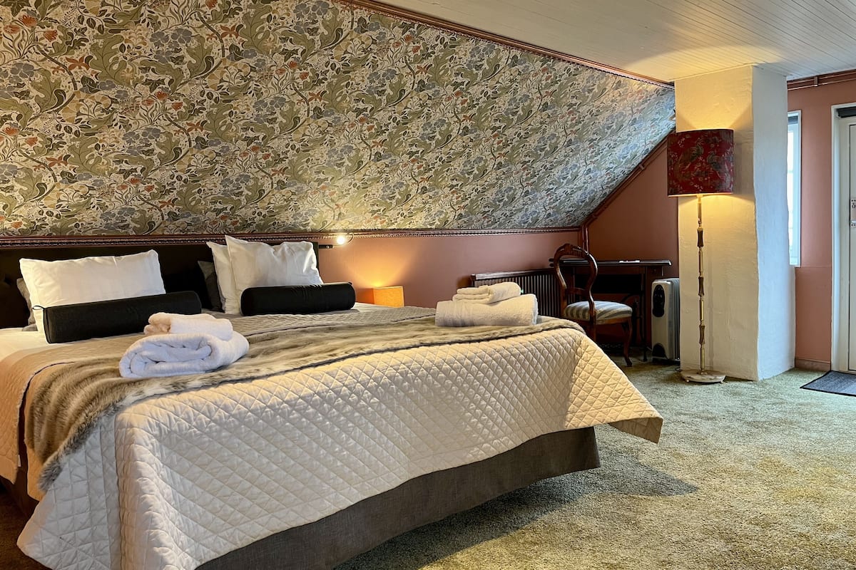 A neatly made bed with a quilted cover, a headboard with a floral wallpaper, a standing lamp, and a radiator.