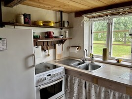 Una cocina con estufa, horno y pileta. Hay repisas sobre la mesada con diversos utensilios de cocina. Se ve una ventana con vista a follaje.