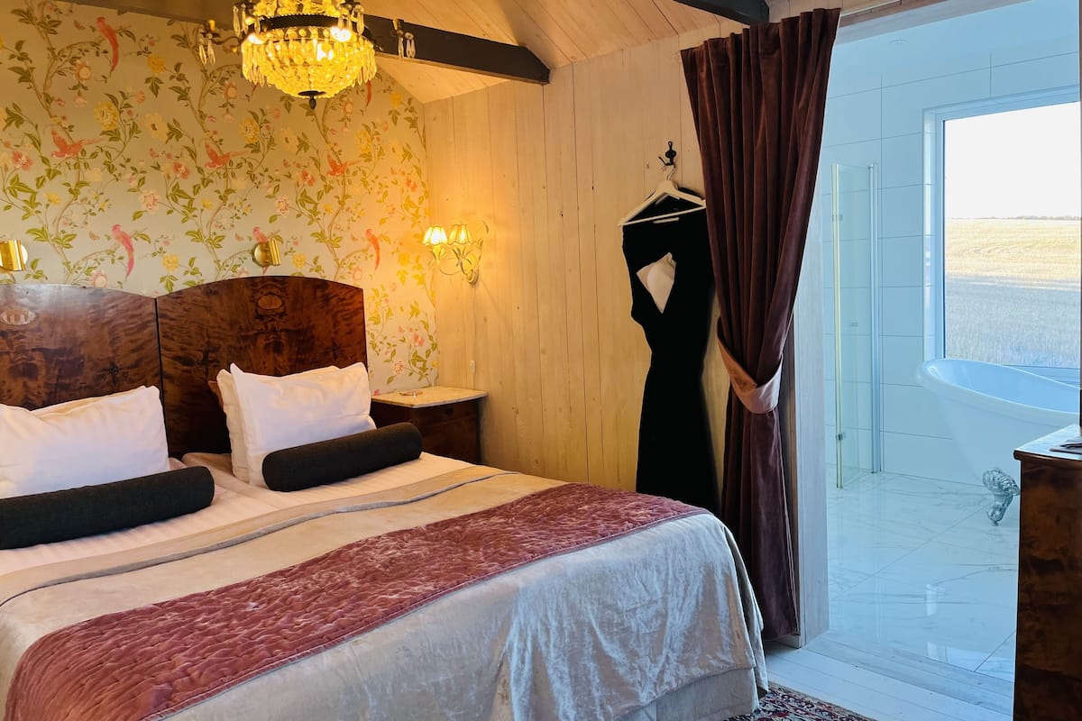 A bedroom with a large bed, a chandelier, a floral wallpaper, a hanging garment, and a view through a window.