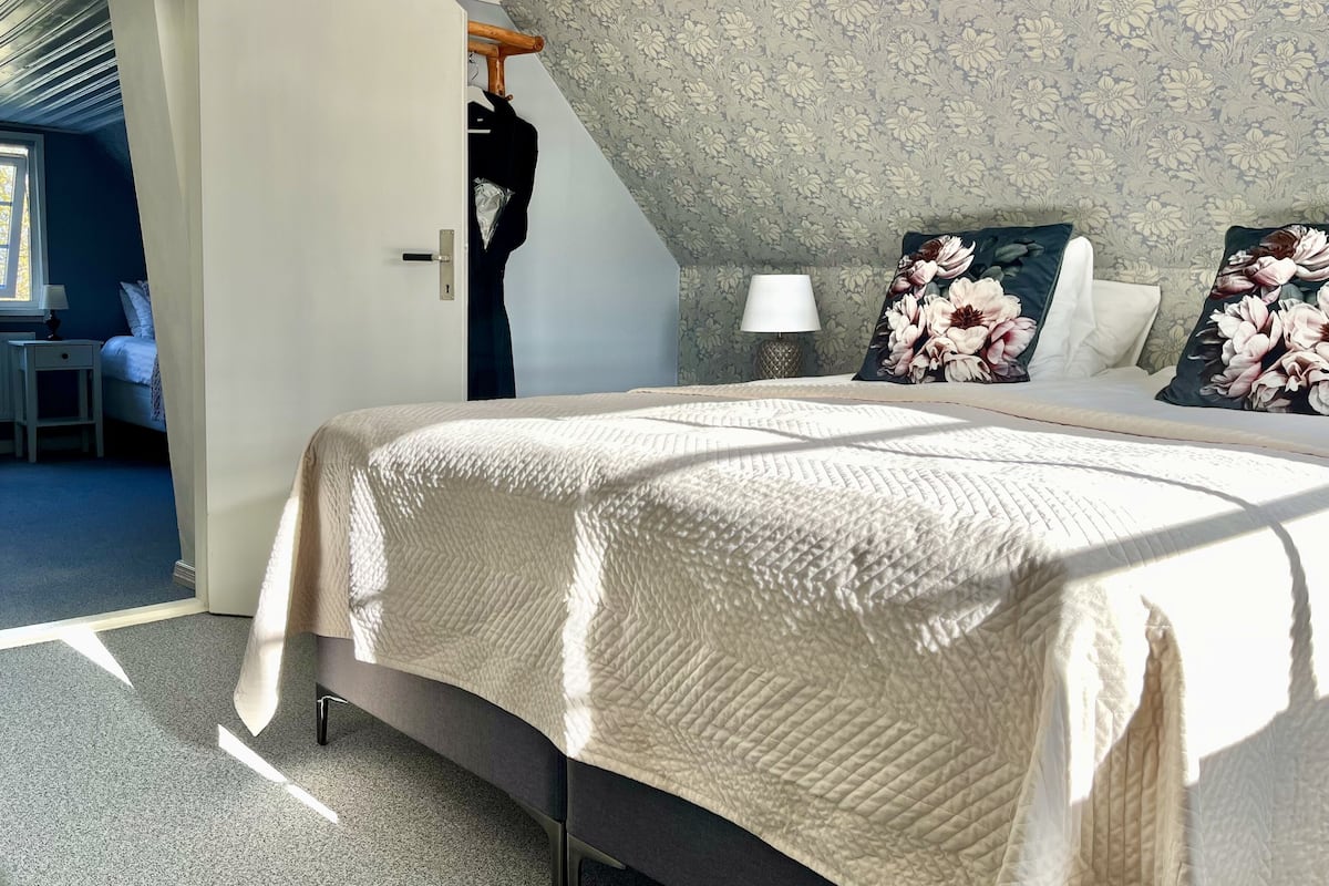 A bedroom with a large bed, floral-patterned pillows, a bedside lamp, and a wardrobe with a coat hanging.