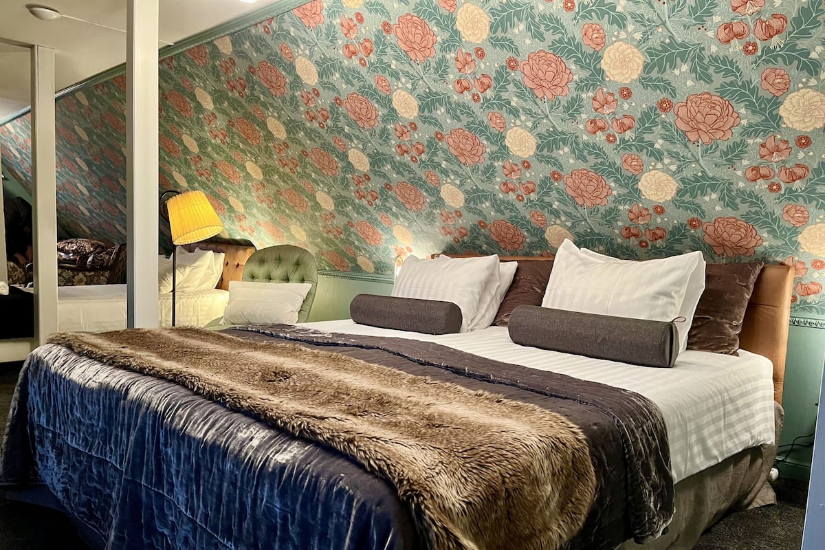 A bedroom with a floral wallpaper, a tufted headboard, a bed with multiple pillows, and a blue blanket.