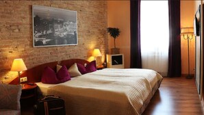 A hotel room with a brick wall, a bed with a white duvet, purple pillows, a nightstand with a lamp, a suitcase, and a framed picture on the wall. - Hotel Berliner Hof Dallgow (Dallgow-Doeberitz)