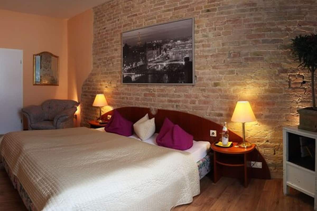 A bedroom with a large bed, two bedside tables with lamps, a chair, a framed picture on the wall, and a brick wall.