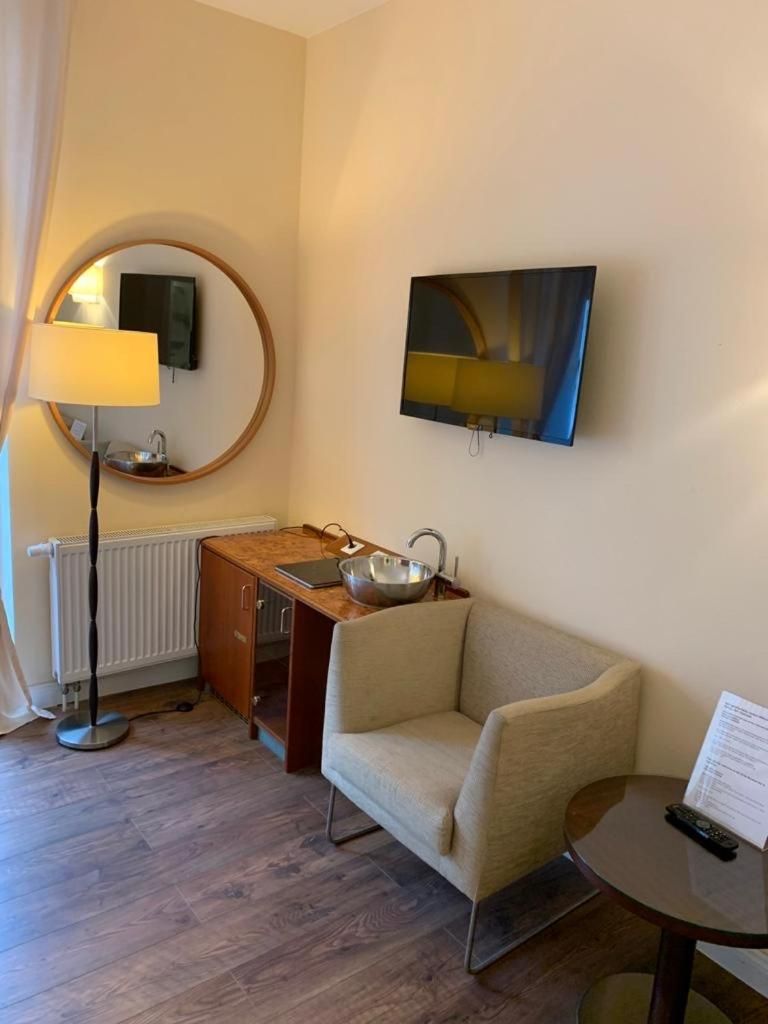 Et hotelværelse med et rundt spejl, et træskrivebord med en skål, en sofa, et lille rundt bord og et TV ophængt på væggen.
