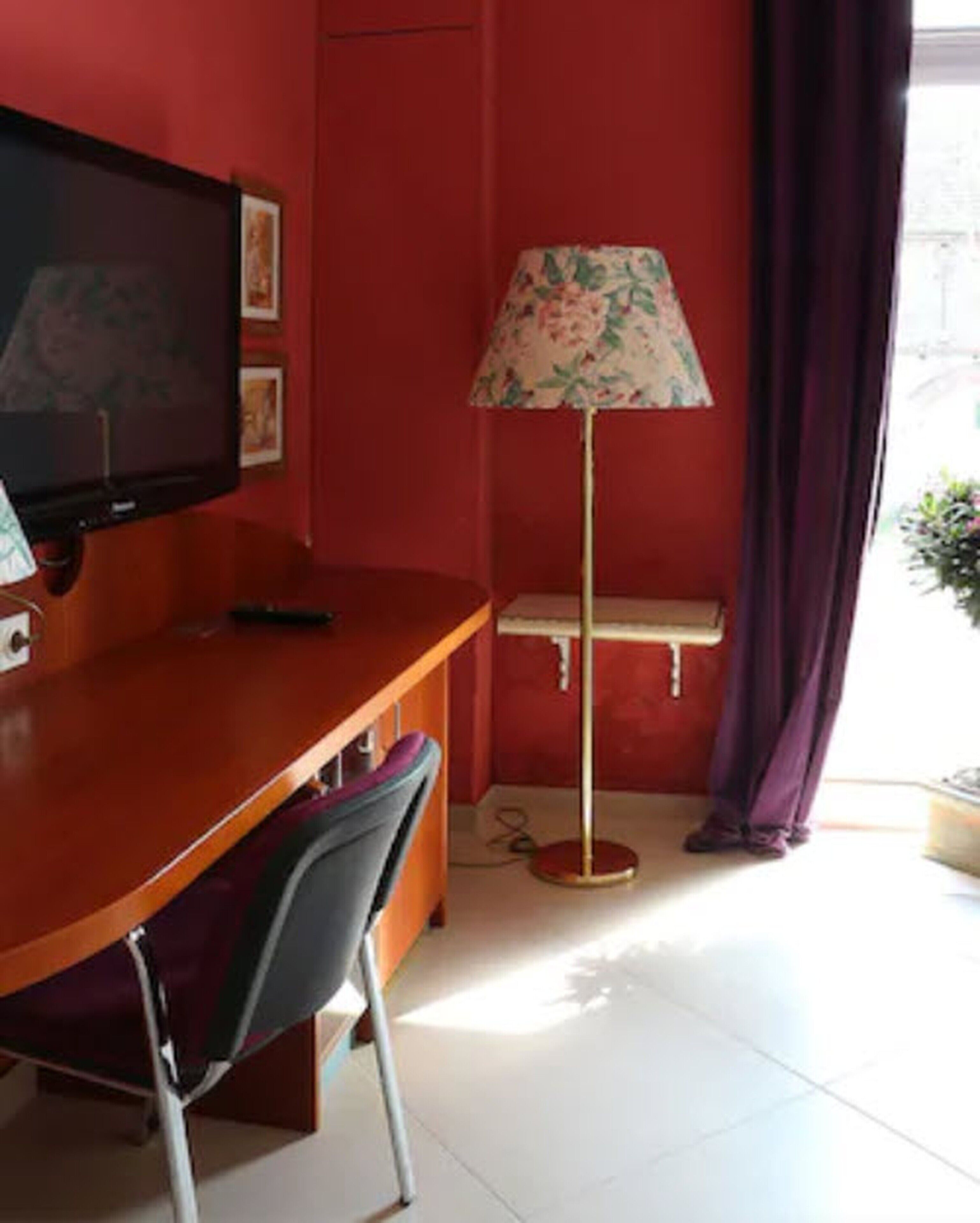 A room with a wooden desk, a chair, a television, a floor lamp, and a window with curtains.