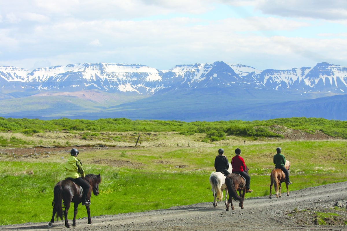 Um grupo de pessoas cavalgando em uma estrada de terra, com uma paisagem montanhosa ao fundo.