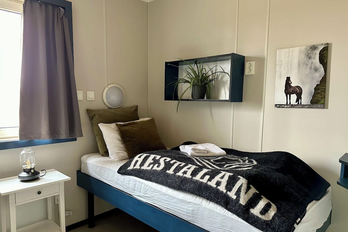 Quarto com cama, mesa de cabeceira com abajur, planta em vaso e prateleira na parede com planta e quadro emoldurado.