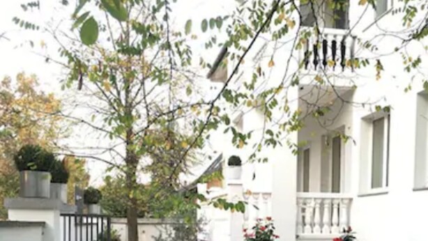 A narrow courtyard with a red carpet leading to a white building with a balcony. There are potted plants and a small tree in the center. - Gatow 183 (Berlin)