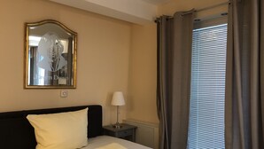 A hotel room with a bed, a nightstand, a lamp, a mirror, and a window with curtains. - Gatow 183 (Berlin)