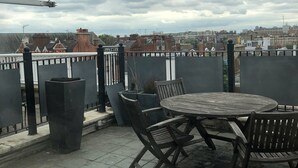 Eine Dachterrasse mit einem runden Holztisch und Stühlen, umgeben von Metallgeländern und Blumentöpfen.