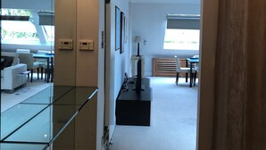Ein modernes Interieur mit Glastrennwand, einem Flachbildfernseher auf einem Standfuß und Blick auf einen Essbereich mit Tageslicht.