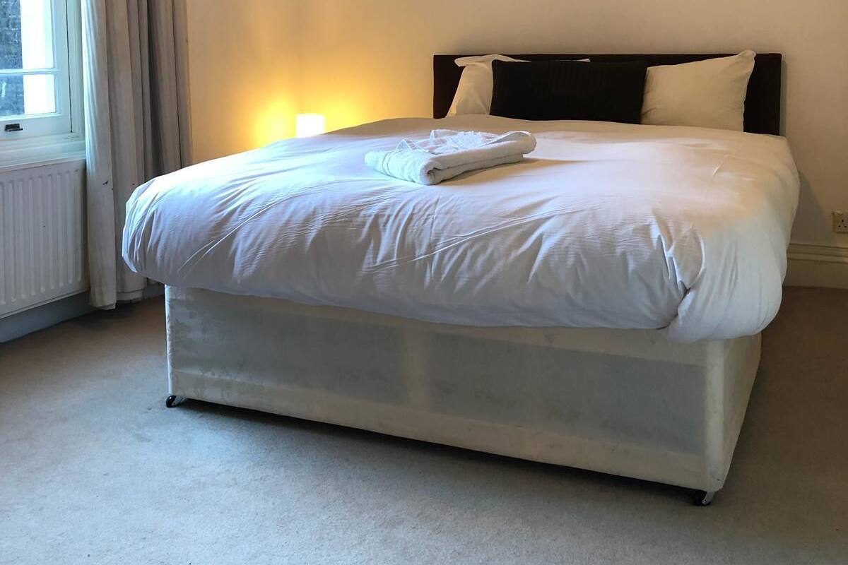 Un lit bien fait, avec du linge de lit blanc, dans une pièce dotée d’une grande fenêtre et d’un radiateur.