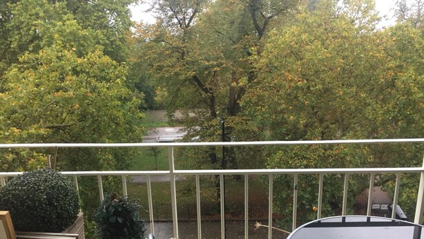 Un balcone con vista su una strada alberata, una ringhiera bianca e un tavolo rotondo.