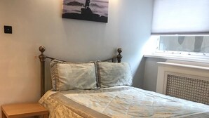Una camera da letto con un letto, un comodino in legno, una lampada a parete, una finestra con persiane e un quadro appeso al muro.
