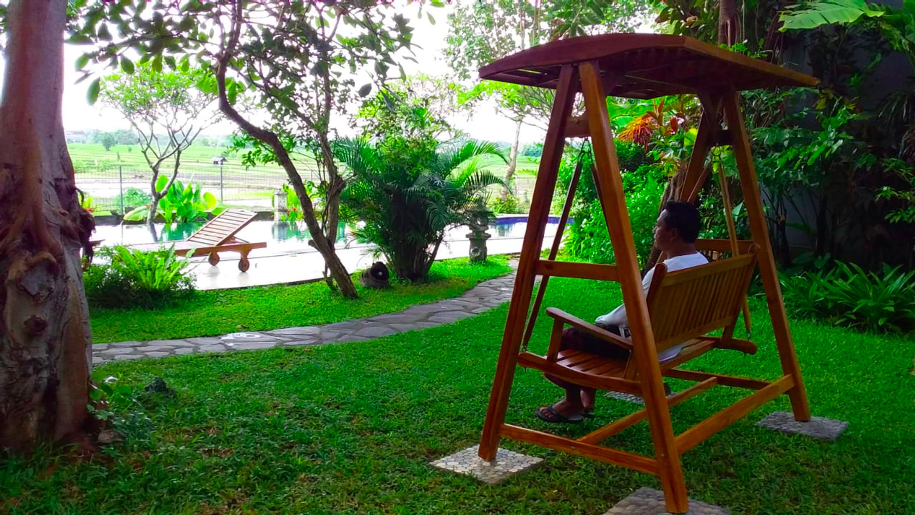A wooden swing chair in a lush garden with a stone pathway and a pool in the background.