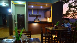Un coin repas avec une table dressée pour deux personnes, une cuisine en arrière-plan et une plante sur la table.