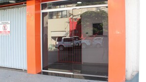 A building with a glass entrance and a red gate, a sign prohibiting parking, and a reflection of a vehicle. - Hotel Flert Tatuape (São Paulo)