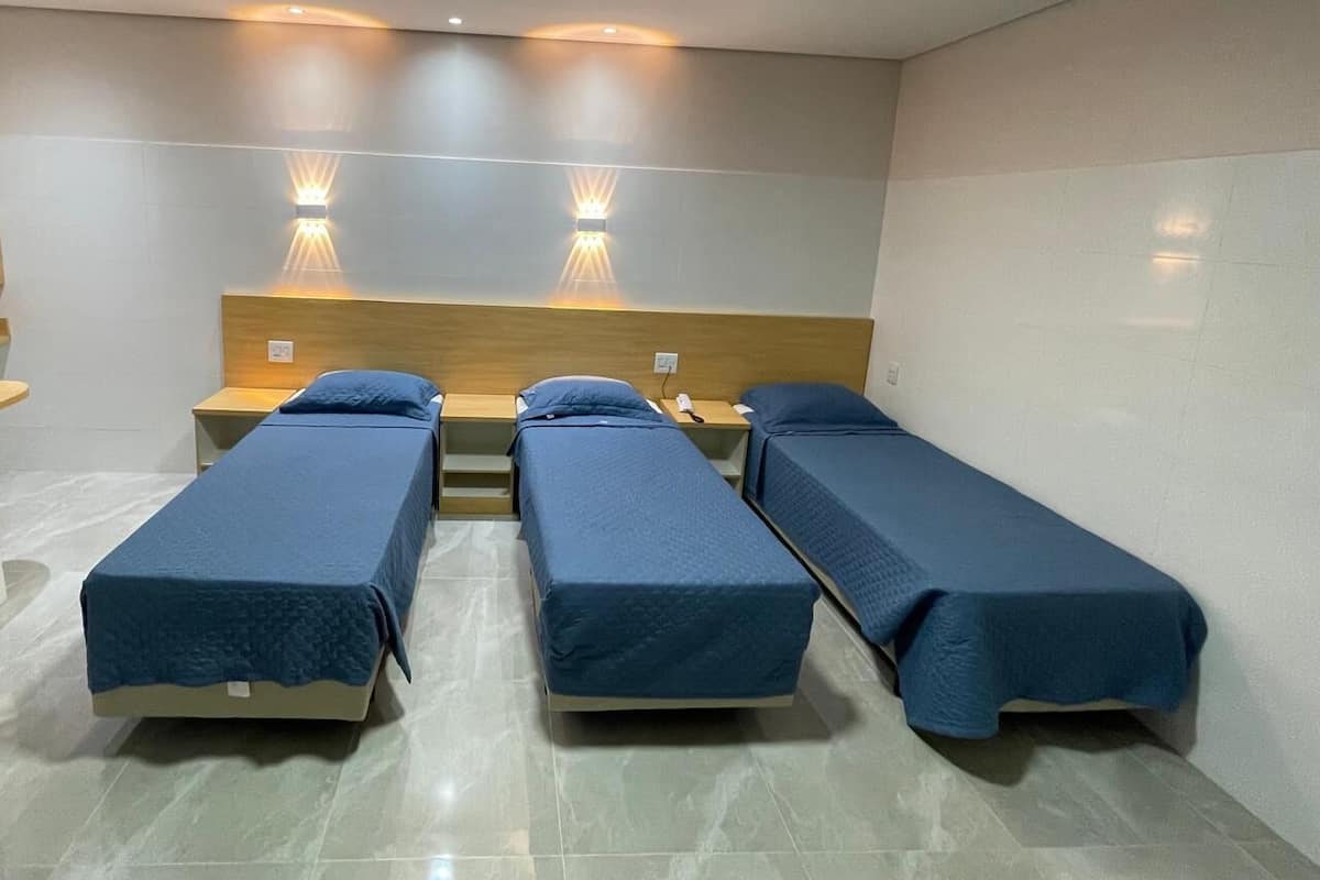A hotel room with three single beds, each with a wooden headboard and a blue bedspread. There are wall-mounted lights above each bed and a ceiling light.