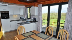 Una cocina con armarios blancos, un microondas y una cafetera. Hay una mesa de comedor con sillas y vistas a un paisaje verde por las ventanas.
