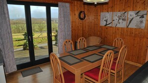 Comedor con paneles de madera, una mesa y sillas, ventanas amplias con vistas al campo y una lámpara fijada a la pared.