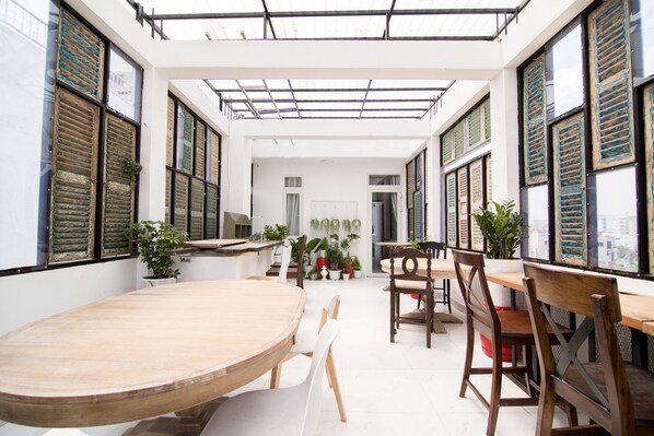 Un vaste espace dédié à la salle à manger, avec une table et des chaises en bois, de grandes fenêtres équipées de volets et des plantes en pot.