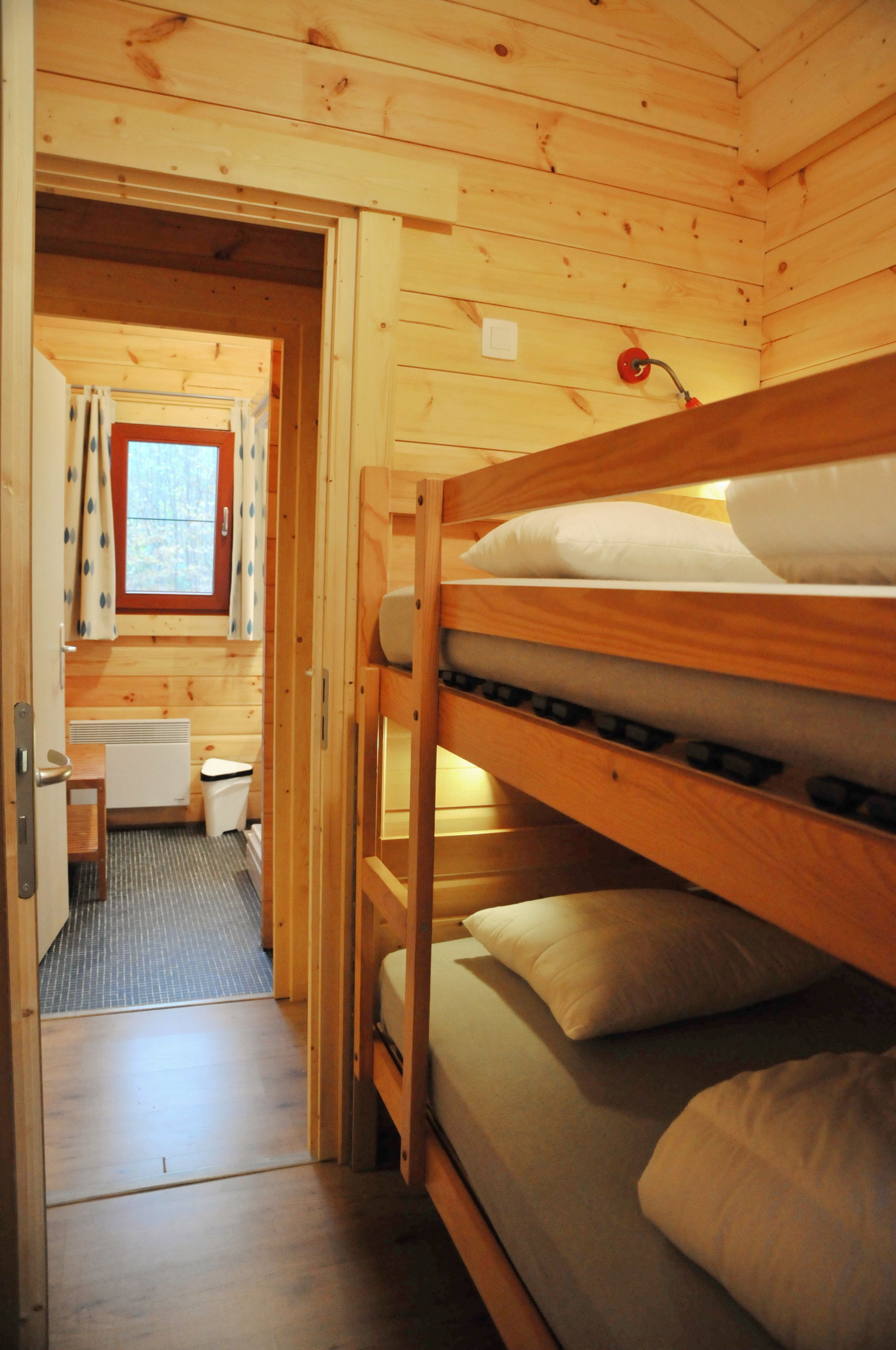 A wooden cabin interior with bunk beds, a small bathroom, and a window with curtains.