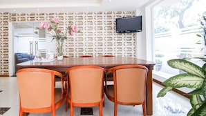 Un comedor con mesa de madera y sillas anaranjadas, un televisor montado en la pared y un ventanal con vistas a zonas verdes.