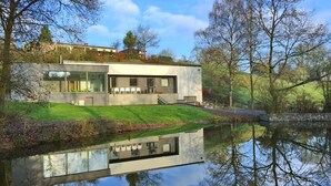 A modern house with a large glass window, a grassy area, and a reflection in the water. - Ard'cube (Somme-Leuze)