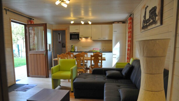 A modern living room with a sofa, chairs, and a coffee table. There is a kitchen area with cabinets and appliances. - Au Fil de l'Eau (Durbuy)