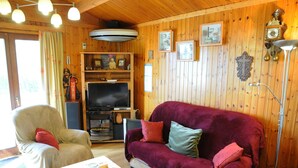 Una habitación con paneles de madera, un televisor, un sofá de terciopelo rojo, un sillón beige y una mesita con controles remotos.