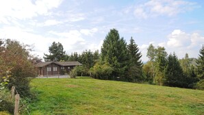 Ein Haus mit geneigtem Dach und einer Veranda, umgeben von einem Rasen und dichten Bäumen.