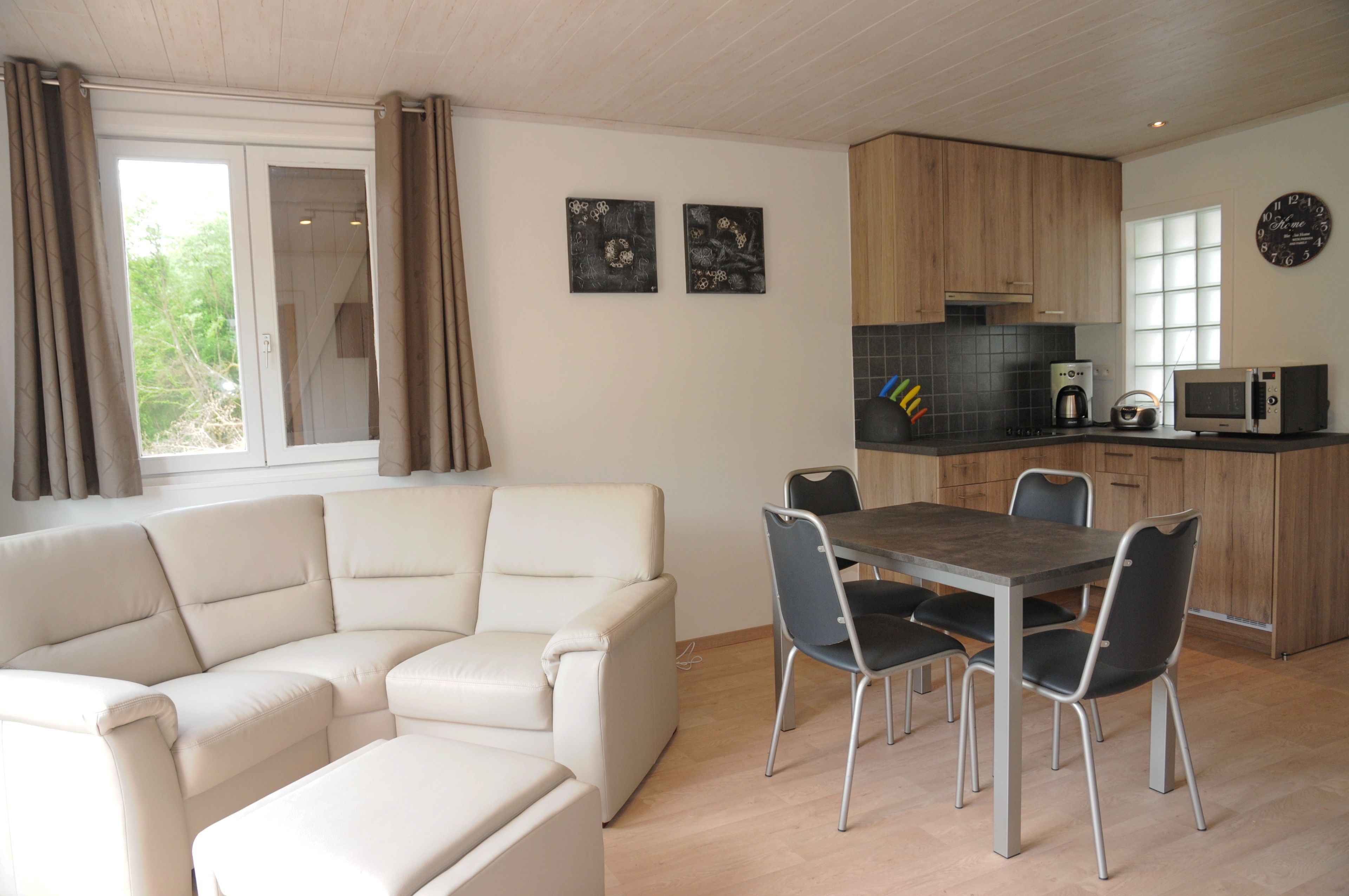 A modern living room with a sectional sofa and a dining table with chairs. There is a kitchen area with wooden cabinets and a microwave. Two abstract paintings are on the wall.