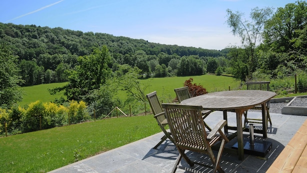 An outdoor patio with a wooden table and chairs, surrounded by a lush green landscape and a forested hill in the background. - Chalet d'aventures (Durbuy)