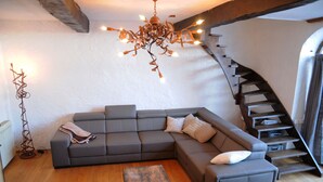 A modern living room with a grey sectional sofa, a wooden coffee table, and a unique chandelier. A spiral staircase is visible to the right. - Chard'heureux (Somme-Leuze)