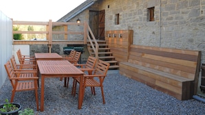 A rustic outdoor seating area with wooden tables and chairs, a stone wall, and a wooden staircase leading to a loft. - Chard'heureux (Somme-Leuze)