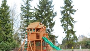 Une cabane en bois avec un toboggan, entourée d’arbres et d’une clôture.