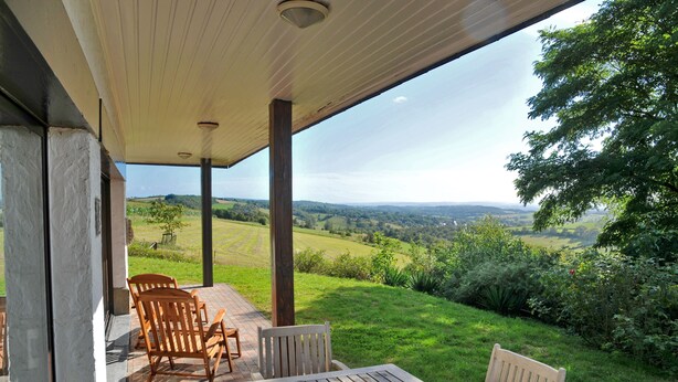A covered patio with wooden furniture and a view of rolling hills. - Chevalerie (Somme-Leuze)