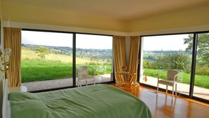 A bedroom with a large bed, two chairs, a small table, and a view of a green landscape through large windows. - Chevalerie (Somme-Leuze)