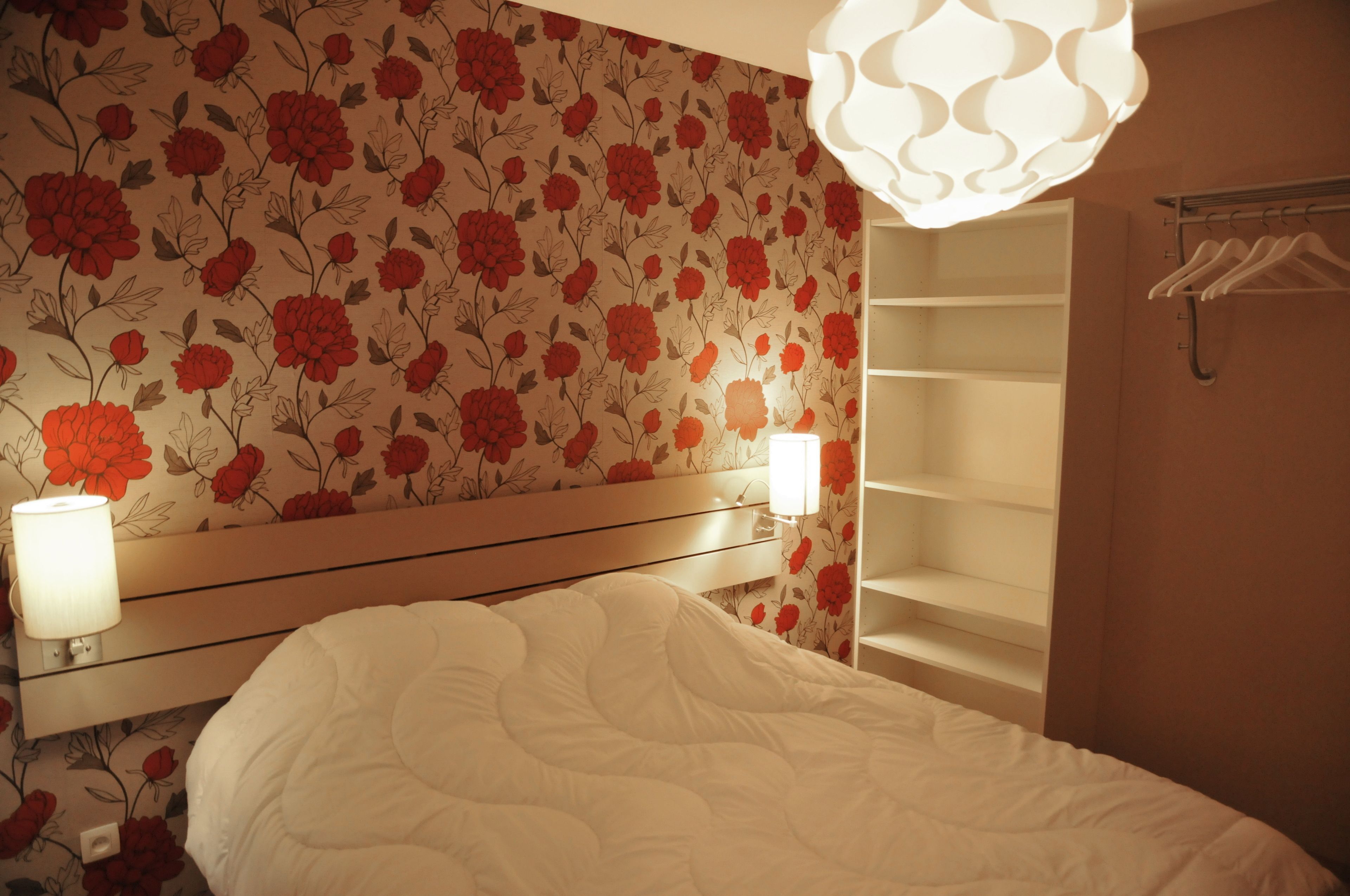 Ein Schlafzimmer mit floraler Tapete, einer Nachttischlampe, einem Bett mit weißer Bettwäsche und einem Kleiderschrank mit Kleiderhaken.