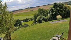 Eine grüne Außenanlage mit einer Bank, zwei Stühlen, einem kleinen Teich und Blick auf sanfte Hügel und Felder.