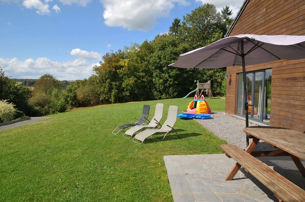 A grassy backyard with two lounge chairs, a picnic table, and a play area with slides and a climbing structure.