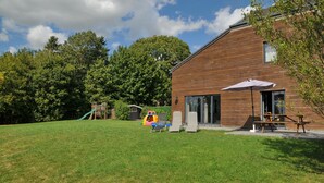 Ein modernes Gebäude mit Holzfassade, einer Terrasse mit Sitzgelegenheiten und einem Spielplatz im Hintergrund.