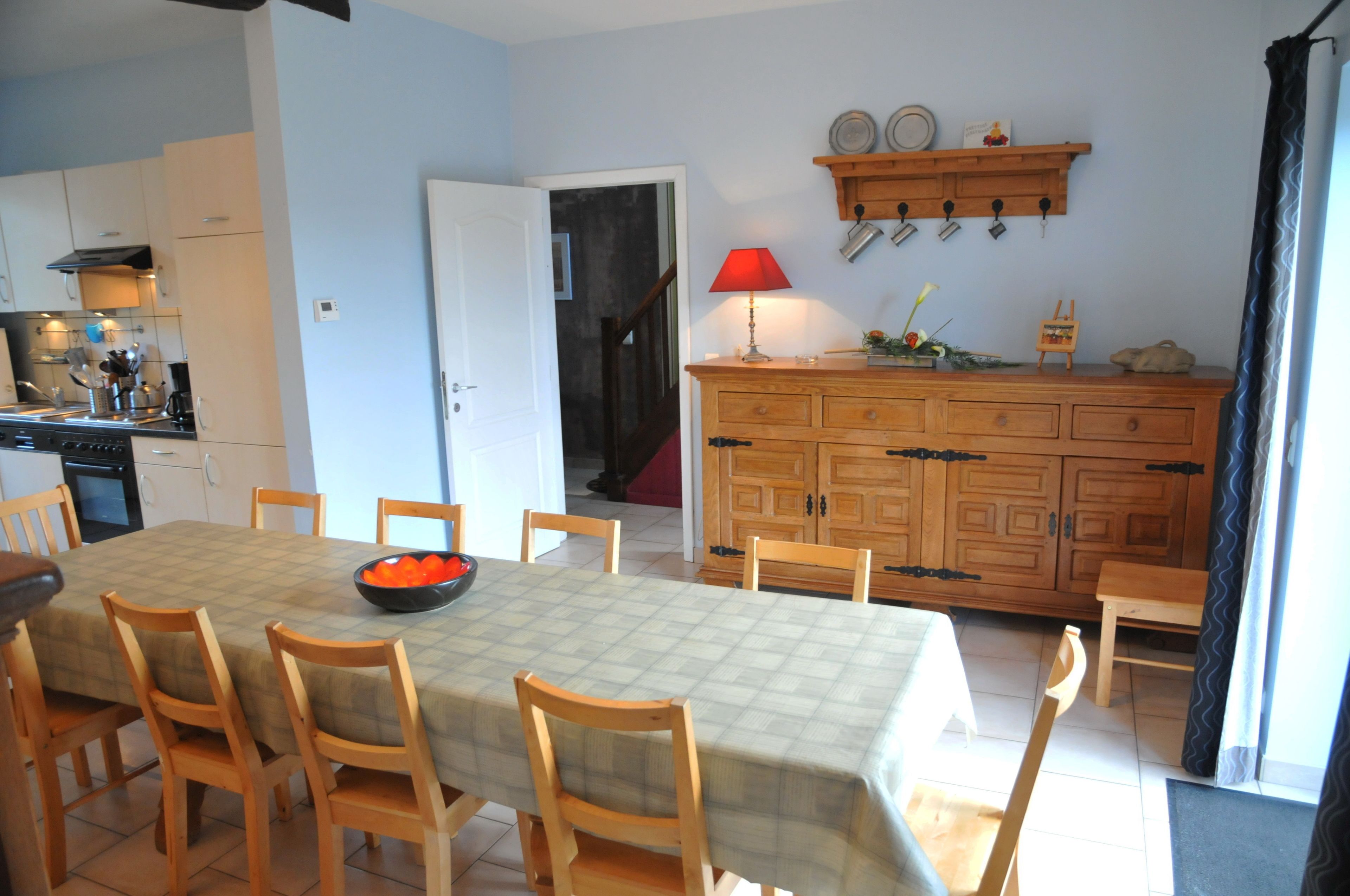 A dining area with a table set for six, wooden chairs, a wooden cabinet with a red lamp, and a kitchen in the background.