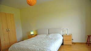 A bedroom with a bed, wooden wardrobe, bedside table with lamp, and a red stool. - Clef des Champs (Somme-Leuze)