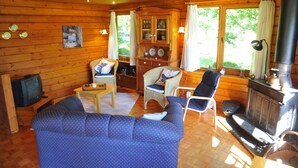 Interior acogedor de una cabaña con un sofá azul, sillas de mimbre, un armario de madera con vajilla, un televisor y un estufa de leña.